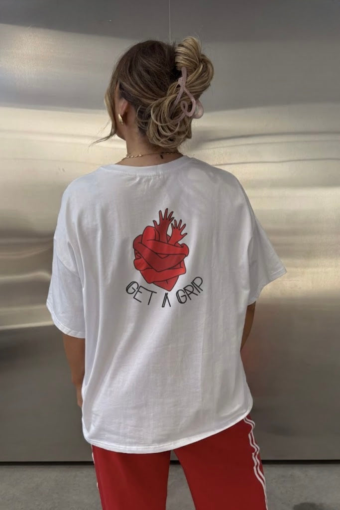 White graphic T-shirt with red heart illustration and “Get A Grip” text printed on the back, worn oversized with red trousers.