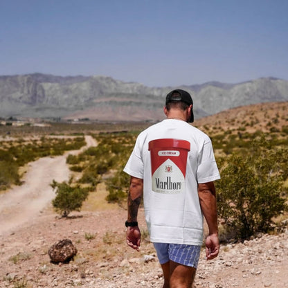 Man wearing a white graphic T-shirt with a red Marlboro-inspired ice cream print on the back, modern streetwear styled outdoors in a desert landscape.